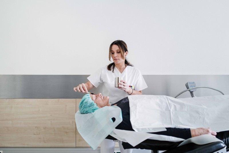 : Eine Patientin liegt auf einer Liege mit geschlossenen Augen und einer OP Haube. Eine Mitarbeiterin reinigt und desinfiziert das Gesicht mit einem großen Wattestäbchen / © Till Findl 