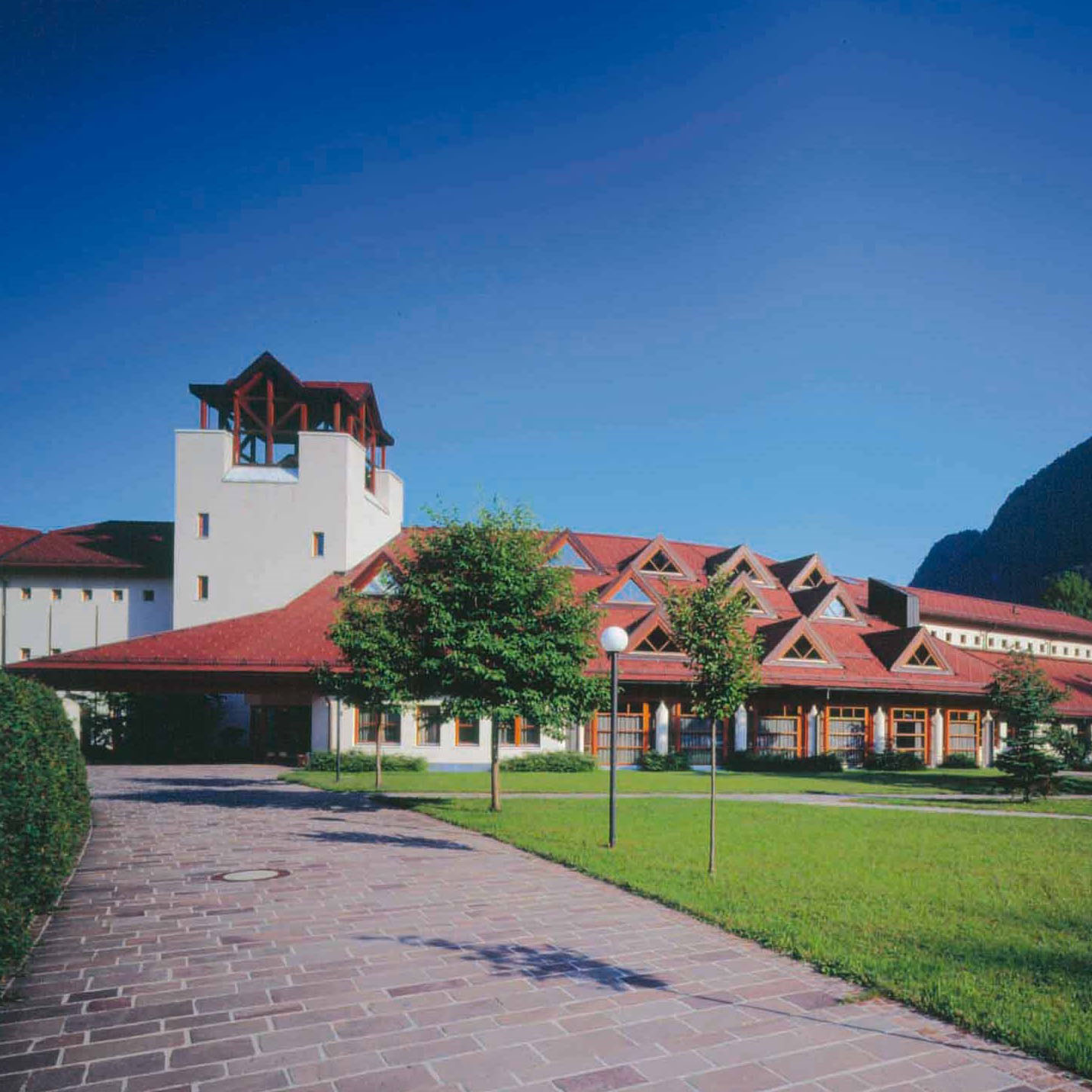 Außenansicht des Gesundheitszentrums: breite gepflasterte Zufahrt mit grüner Hecke links und rechts Bäumen, Wiese und Wegen; Das Gebäude ist bungalowartig mit Dreiecksgaupen in drei Reihen am Dach; Auffällig ist ein Turmabau hinter dem Eingangsbereich.