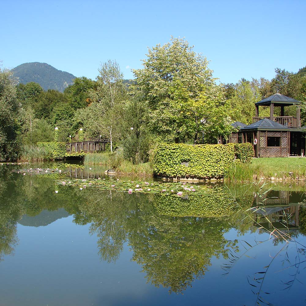 Wunderschöne Gartenanlage des Gesundheitszentrums: im Vordergrund ein Teich mit Seerosen, am Ufer beginnt die Gartenanlage mit kleinen Holzpavillons, Wiesen, Bäumen und Sträuchern. Eine Holzbrücke führt über den Deichzufluss.