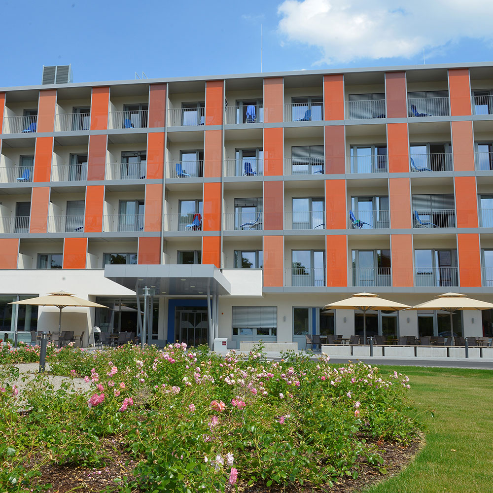 Außenansicht Gesundheitszentrum Peterhof; Vierstöckiger Flachdachbau mit Sicht auf Balkone mit orangen Trennwänden; links und rechts vor dem überdachten Eingang stehen Sitzgruppen mit Sonnenschirmen; im Vordergrund wachsen Rosen und eine Wiese;