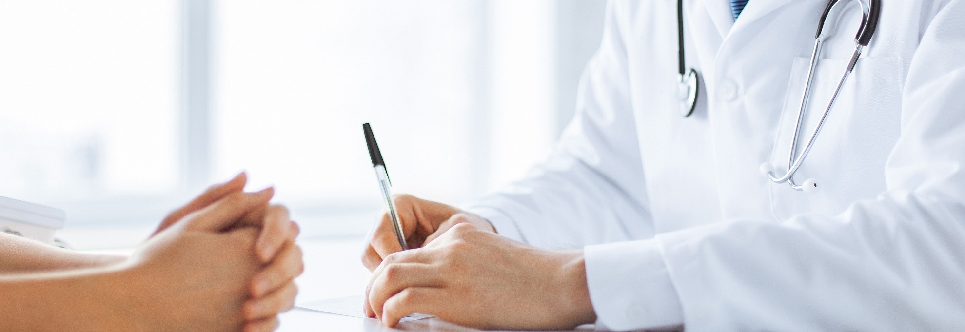 Arzt schreibt mit einem Stift auf ein Blatt Papier. Gegenüber sitzt eine Patientin.