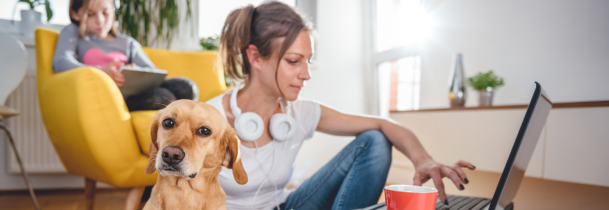 Frau mit Kopfhörern um den Hals arbeitet am Laptop. Im Vordergrund ein hellbrauner Hund. Im Hintergrund ein Mädchen auf einem Polstersessel, das ein Tablet bedient.