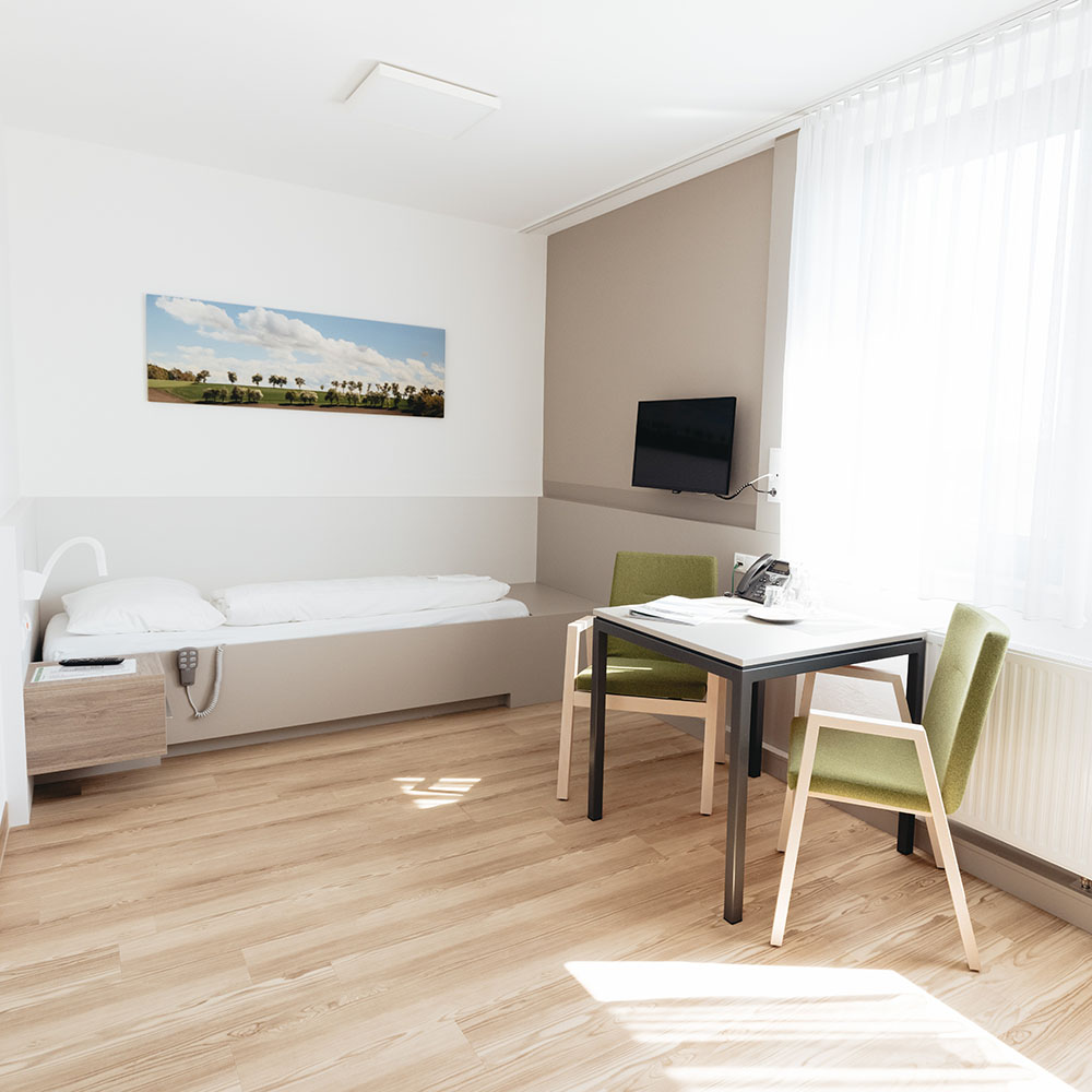 Helles Einbettzimmer mit Parkettboden; am Fußende des Bettes hängt ein Fernseher an der Wand, neben dem Bett vor einem Fenster zwei grüne Sesseln und ein Tisch mit Telefon; gegenüberliegend neben dem Bett ein Nachtkästchen 