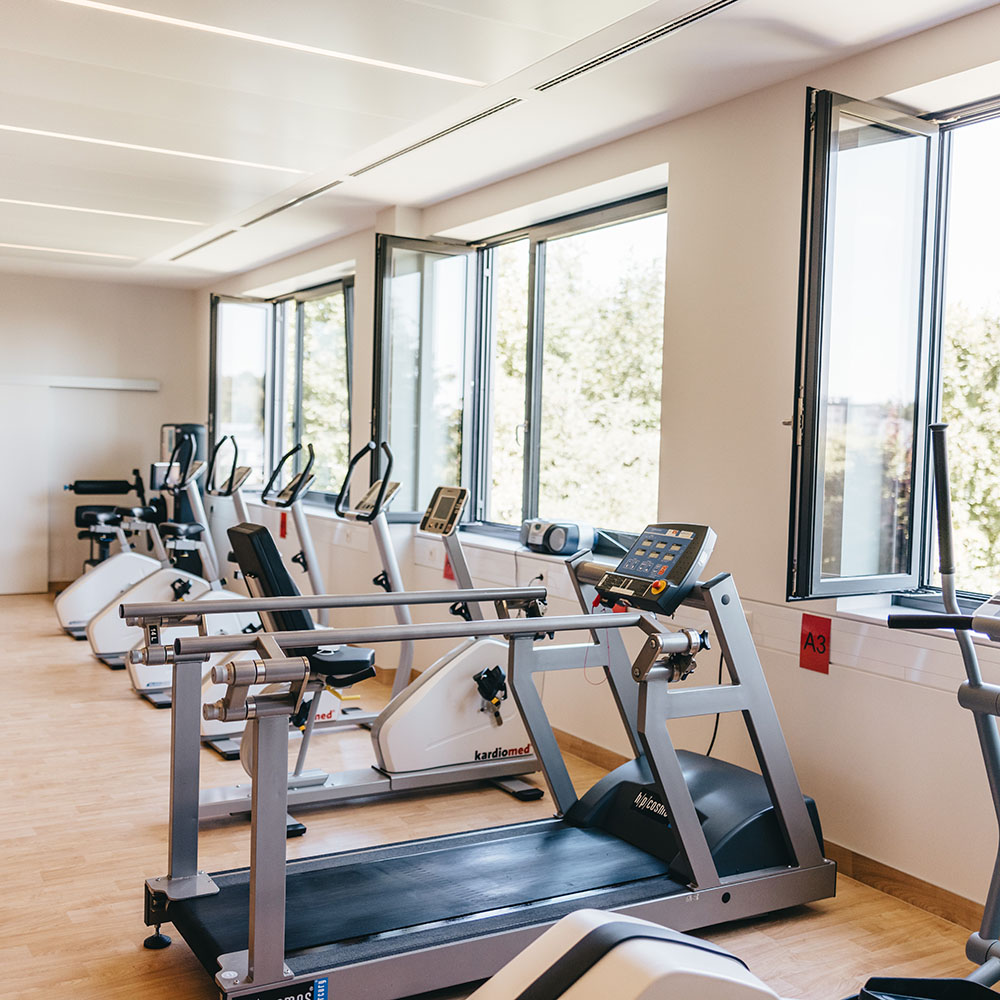 Fitnessraum im Gesundheitszentrum: Ein Laufband und verschiedene Ergometer stehen in einer Reihe vor einer breiten Fensterfront mit Blick ins Grüne. 