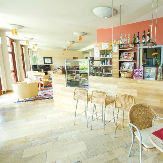 Geräumige Cafeteria mit Barhockern vor einem Tresen mit Snacks und Getränken. Im Hintergund bequeme Polstersessel an kleinen Tischchen.
