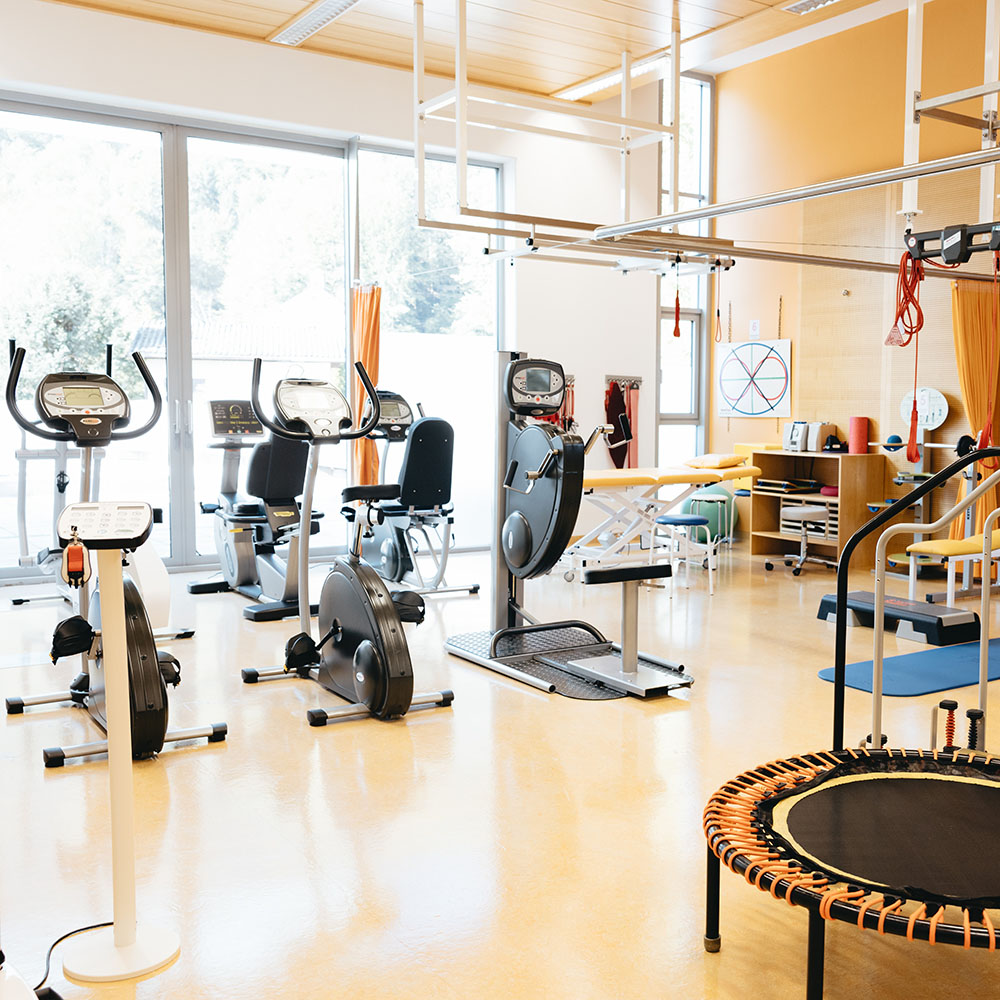 Großer Trainingstherapieraum im Gesundheitszentrum; breite hohe Fensterfront mit Blick ins Grüne, davor stehen in zwei Reihen jeweils drei Trainingsgeräte, rechts vorne ein Trampolin, rechts hinten im Bild ein mit Vorhang abtrennbarer Bereich mit Liege;