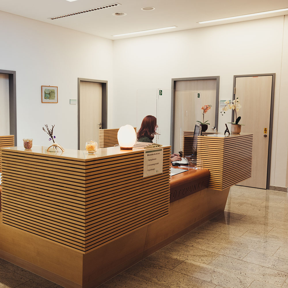 Anmeldebereich für Therapien: große viereckige Rezeption mit Covid-Schutz-Glas vor dem Anmeldebereich; dahinter sitzt eine Frau und arbeitet am Computer; auf der Theke stehen Orchideen und andere Accsessoires; Türen zu Therapieräumen im Hintergrund;