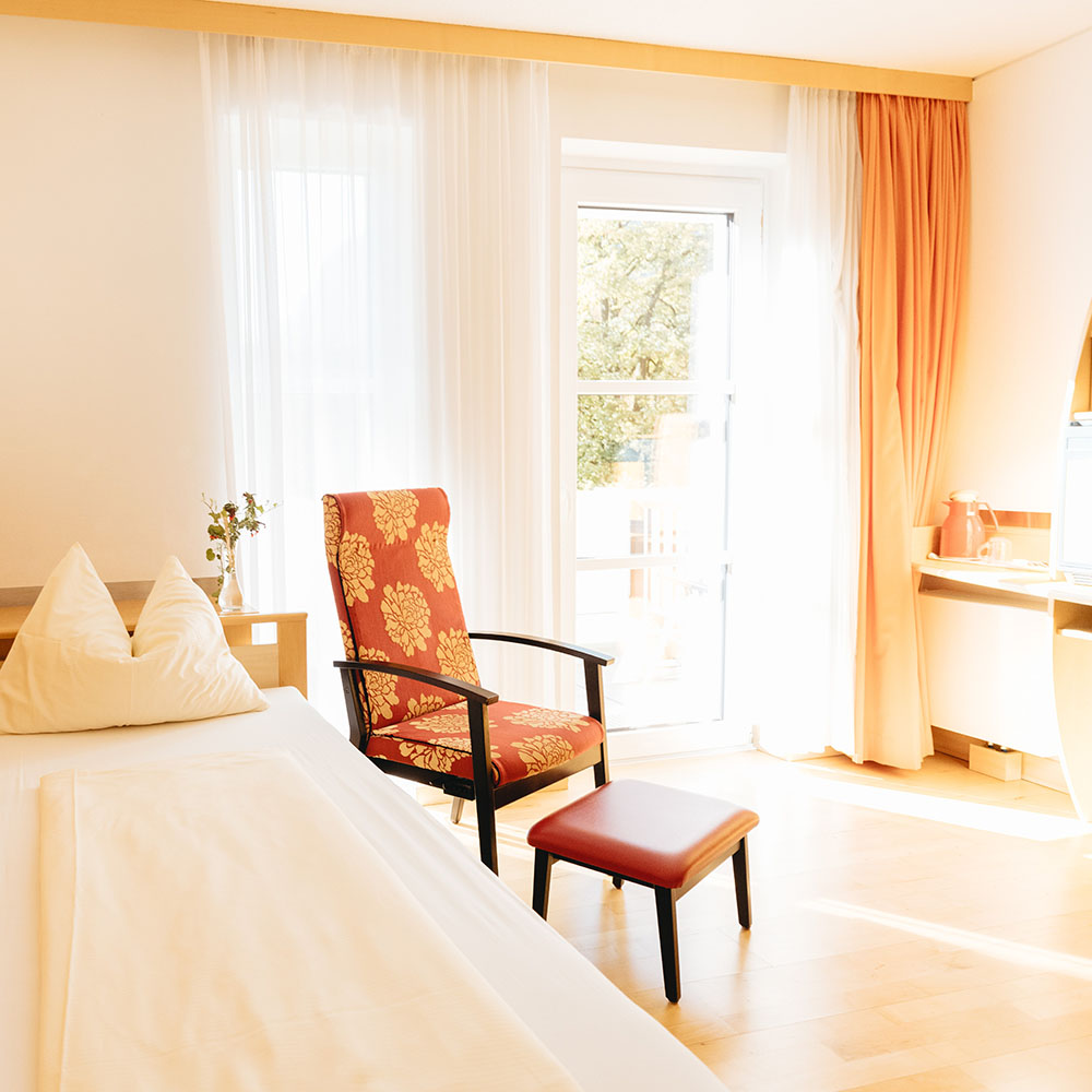 Helles sonniges Einzelzimmer mit Balkon und Blick auf Bäume. Vor dem Bett steht ein orange tapezierter Sessel mit Hocker, ebenfalls hellorange sind die Vorhangseitenteile.
