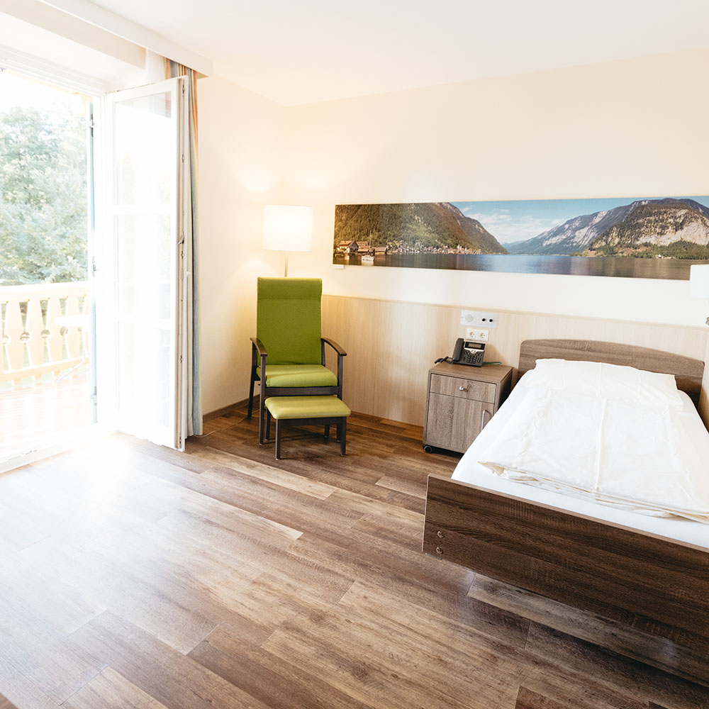 Einzellbettzimmer mit dunklem Parkettboden und ebensolchen Möbeln, helle Wände, grüner Sessel und Blick auf den Balkon mit Aussicht ins Grüne.