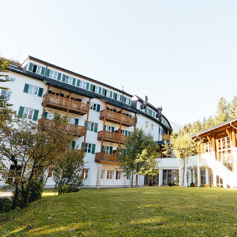 Rückseitige Außenansicht des Gesundheitszentrums Hanuschhof. Fenster und Holzbalkone mit Sicht auf eine Wiese mit Bäumen.