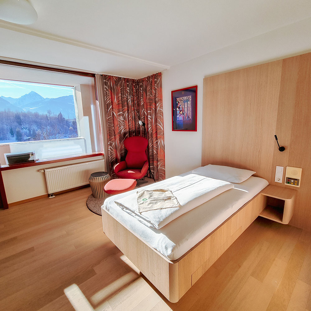 Helles Zimmer mit einem Bett, einem Sessel, Holzfußboden und einem fenster mit Blick auf Wald und Berge.